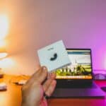 Hand holding an AirTag box in front of a laptop on a cozy desk setup.